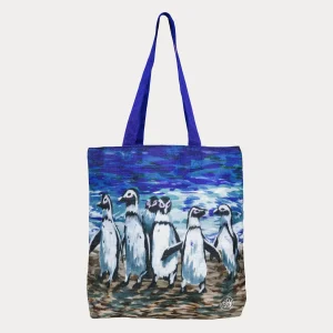 A tote bag with blue handles featuring a colorful painting of African penguins standing together on a sandy beach with a blue ocean background.